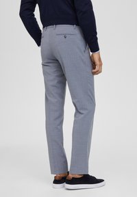 Pantaloni grigi con un tessuto testurizzato, taglio slim e tasche posteriori con bottone. Abbinati a scarpe scure con suola bianca e dettagli marroni.