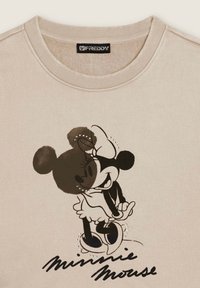 Sweatshirt beige avec un graphique noir et blanc de Minnie Mouse, accompagné d'un texte manuscrit "Minnie Mouse" en dessous, et un design à col rond avec côtes.