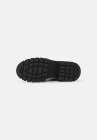 Black rubber shoe sole with deep tread pattern, featuring a rounded toe and a textured surface for enhanced grip.