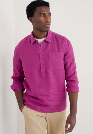 Camisa de lino fucsia con cuello, cuello abotonado, un bolsillo en el pecho y mangas remangadas, combinada con pantalones beige.