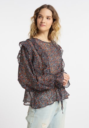 Femme aux cheveux ondulés portant un chemisier floral transparent avec des volants et un jean déchiré bleu clair, debout devant un fond uni.