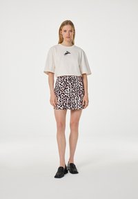 Vrouw in een lichtbeige oversized T-shirt met een klein zwart graphic en een roze en zwart gestippelde minirok, staande op een witte achtergrond.