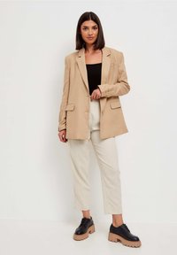 Blazer oversized beige, design ajusté, deux poches avant, associé à un pantalon crème et des chaussures à plateforme noires épaisses. Tissu lisse, coupe structurée.