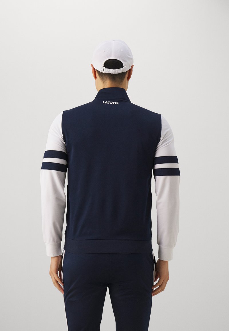 Veste de sport bleu marine avec des manches blanches, arborant trois bandes bleu marine sur chaque bras et un col haut. Logo Lacoste dans le cou.