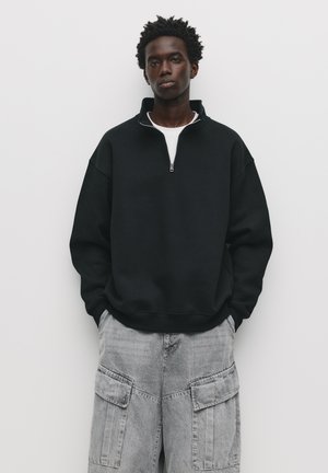 Sort sweatshirt med lynlås ved kraven og lange ærmer, parret med grå cargo-bukser med flere sidelommer.