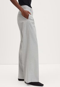 Pantaloni grigi chiari a gamba larga con una texture liscia, tasche laterali e una vita aderente, abbinati a scarpe nere con lacci decorativi.