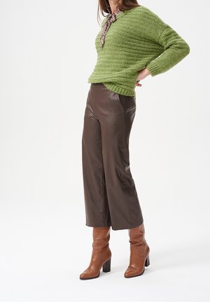 Kvinde iført grøn strikket trøje, brune beskårne læderbukser, brune støvler med hæl og leopardmønstret halstørklæde, stående med hånden på hoften.