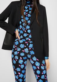 Blazer entallado negro combinado con un cuello alto negro con estampado floral y leggings. El patrón floral presenta flores azules con hojas rosas sobre un fondo negro.