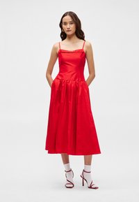 Κόκκινο σατέν φόρεμα με εφαρμοστό κορσέ, λεπτές τιράντες και φούστα φουσκωτή, μέχρι το γόνατο. Το μοντέλο φοράει κόκκινα τακούνια τα οποία ταιριάζουν και λευκές κάλτσες.