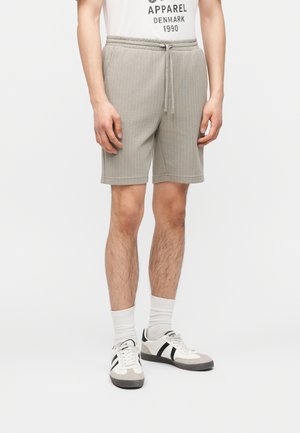 Man draagt beige geribde short met trekkoord, witte sokken en witte sneakers met zwarte strepen, staand tegen een effen achtergrond.