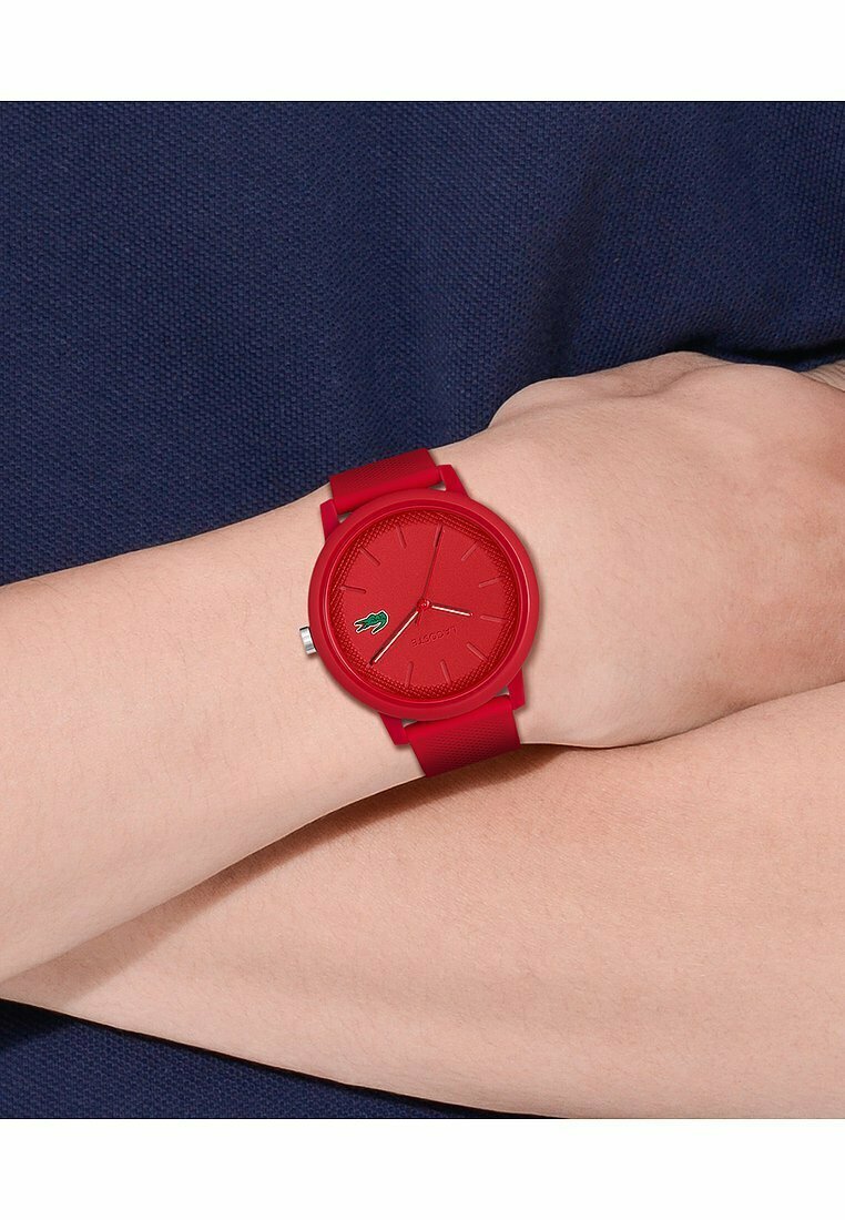 Orologio in silicone rosso con cassa rotonda, quadrante testurizzato e marker delle ore minimalisti. Presenta un dettaglio del logo verde sul quadrante.