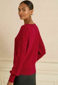 Punainen neulepaita pitkillä, puffimaisilla hihoilla, joissa on timanttikuvioita. Nyöritetty kaula-aukko ja ribatattu helma, käytetty mustan hameen päällä.