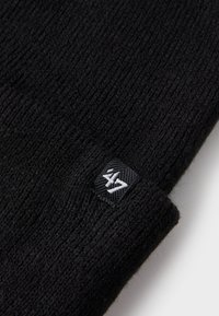 Sort strikket hue med ribbet tekstur, der har et lille firkantet mærke med et hvidt logo på siden. Blødt materiale for varme.