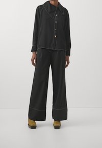 Černá košile na knoflíky s velkým límcem a bílým prošíváním, kombinovaná s černými kalhotami s širokými nohavicemi. Oblečeno s hnědými chunky podpatky.