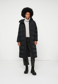 Kvinne med afro som har på seg en lang svart puffjakke, lys grå turtleneck genser, svarte jeans og svarte snørebots, står mot hvit bakgrunn.