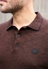 Homme portant un polo marron texturé avec trois boutons et un petit logo rond noir sur la poitrine, montrant le côté du cou et la barbe.