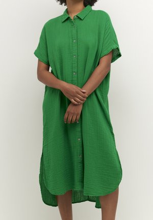 Personne portant une robe verte ample à manches courtes avec des boutons et un col, debout les mains croisées sur le milieu du corps.