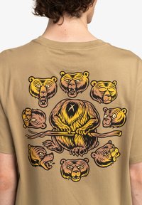 Tan t-shirt featuring a colorful graphic of a bear sitting on a branch, surrounded by various bear head illustrations in shades of brown and yellow.