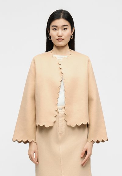 Vrouw draagt een beige jasje met geschulpte rand over een witte top en bijpassende beige rok, staand tegen een effen achtergrond.