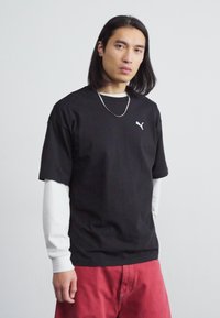 Tricou negru cu mânecă scurtă, cu un logo alb Puma, suprapus peste un cămașă albă cu mânecă lungă. Modelul poartă pantaloni roșii.