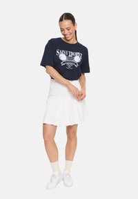 T-shirt bleu marine avec impression graphique blanche, associé à une jupe blanche plissée. Des baskets blanches et des chaussettes claires complètent la tenue.