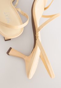 Sandales à talons hauts nude avec brides et finition brillante ; présentent un talon effilé et une semelle intérieure rembourrée. Logo de la marque visible sur la semelle.