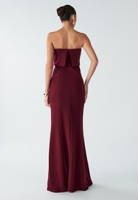 Burgundská bezramínková róba s přiléhavým střihem, texturovanou látkou a detailem zadního skládání. Hladký povrch a skrytý zip na zadní straně.