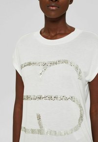 Weißes T-Shirt mit kurzen Ärmeln, das ein glitzerndes silbernes Logo-Design mit Pailletten und einer weichen, leichten Textur aufweist.