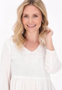 Fehér, hosszú ujjú blúz V-nyakkal, díszített hímzéssel és ráncolt derékkal. Puha, könnyű anyag, finom textúrával.