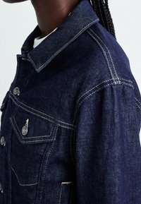 Mörkblå jeansjacka med klassisk krage, frampocket med knäppning, samt kontrasterande sömmar längs kanterna.