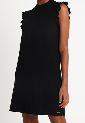 Robe noire sans manches avec un col montant et des détails à volants sur les épaules, portée par une personne debout avec un bras détendu.