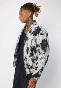 Veste bomber noire et blanche avec un motif abstrait ressemblant à de la peinture, fermeture éclair sur le devant, poignets côtelés et coupe décontractée.