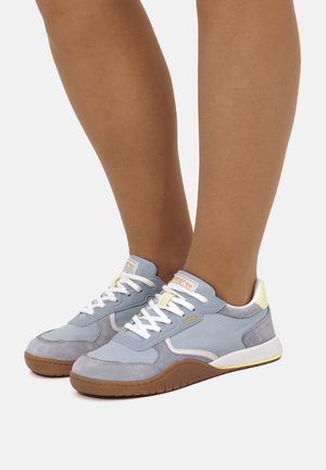 Sneakers azzurro chiaro e grigio con lacci, suole bianche e suole esterne marroni, indossate a piedi nudi su uno sfondo semplice.