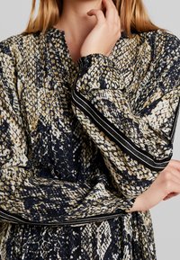 Šaty s dlouhým rukávem v modrém, béžovém a černém hadím vzoru. Mají výstřih do V a pruhované detaily na rukávech.