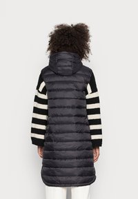 Czarna, pikowana kamizelka z kapturem. Rękawy wykonane z czarno-kremowego, prążkowanego dzianego materiału. Długość sięga do połowy ud.