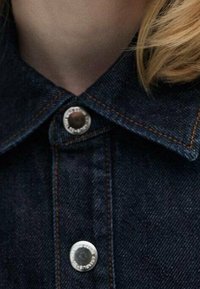 Primer plano de una chaqueta de denim oscuro con un collar puntiagudo y dos botones a presión metálicos, con costuras en naranja contrastante a lo largo de las costuras.