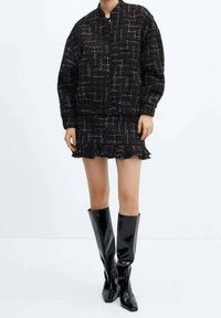 Veste bomber en tweed noir avec un motif blanc, associée à une jupe à ourlet à franges noire et des bottes hautes en vernis brillantes. Texture lisse.