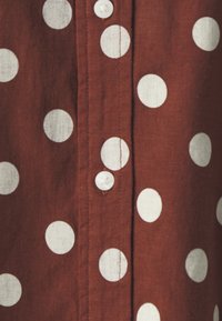 Camisa de tela marrón con puntos blancos, cierre de botones y un acabado texturizado. Los puntos están distribuidos de manera uniforme en la parte delantera.