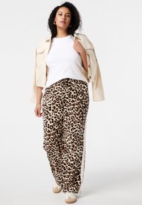 Leopardenmuster weit geschnittene Hose mit elastischem Bund, kombiniert mit einem weißen T-Shirt und einer hellbeige übergroßen Jacke. Sneakers runden den Look ab.