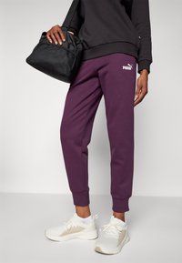 Lila joggingbyxor med vitt Puma-logga, tillsammans med vita sportskor. En svart väska med en texturerad yta hålls i ena handen.