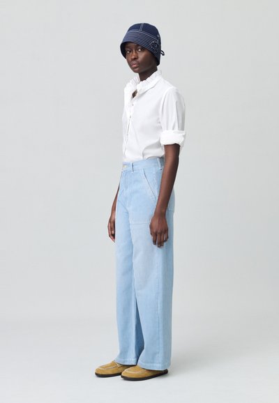 Chemise blanche à volants, pantalons larges rayés bleu clair, chapeau seau marine, et chaussures à enfiler beiges. Look décontracté et ample.