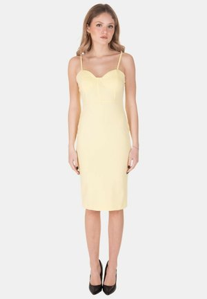 Donna con capelli mossi che indossa un vestito aderente di colore giallo pallido, lungo fino al ginocchio, con bretelle sottili e tacchi neri appuntiti, in piedi davanti a uno sfondo bianco.