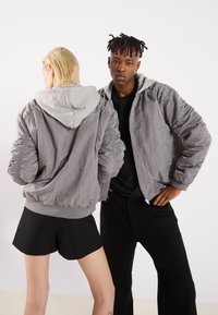 Grauer Bomberjacke mit leichter grauer Kapuze, gerippten Bündchen und einer leicht zerknitterten Textur. Modelle tragen Shorts und schwarze Hosen.