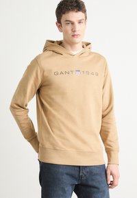 Felpa beige con tasca a canguro, cappuccio con coulisse e logo stampato "GANT 1949" in blu scuro. Morbido misto cotone, vestibilità classica.