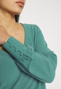 Žena v tyrkysové blůze s dlouhými rukávy a knoflíky potaženými látkou na manžetách, ruka se dotýká klíční kosti, neutrální pozadí.