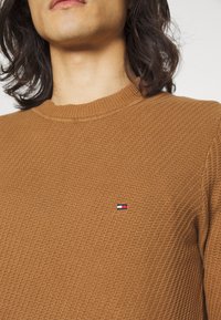 Maglione marrone a trama con scollo rotondo, caratterizzato da un piccolo logo rosso, bianco e blu sulla parte sinistra del petto.