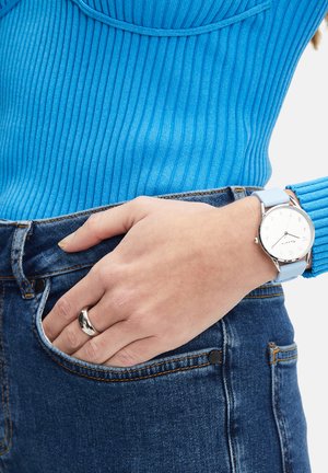Zilveren polshorloge met een witte wijzerplaat en een lichtblauwe siliconen strap, gedragen door een hand die op donkere denim jeans rust. Blauwe geribde top zichtbaar.