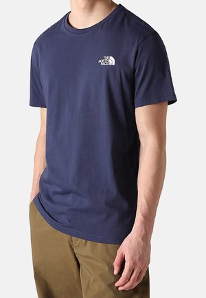 Mand iført en navyblå The North Face T-shirt med korte ærmer og brune bukser, stående med den ene hånd afslappet ned langs siden.