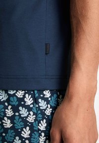 Dunkelblaues Baumwoll-T-Shirt mit genähtem Saum und einem kleinen Etikett. Kombiniert mit gemusterten Shorts in Teal- und Weißtönen mit Blattdesign.