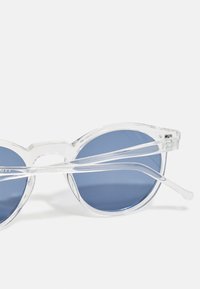 Lunettes de soleil en plastique transparent avec des verres ronds bleus et un design audacieux et minimaliste. La monture présente des bords lisses et des branches fines.